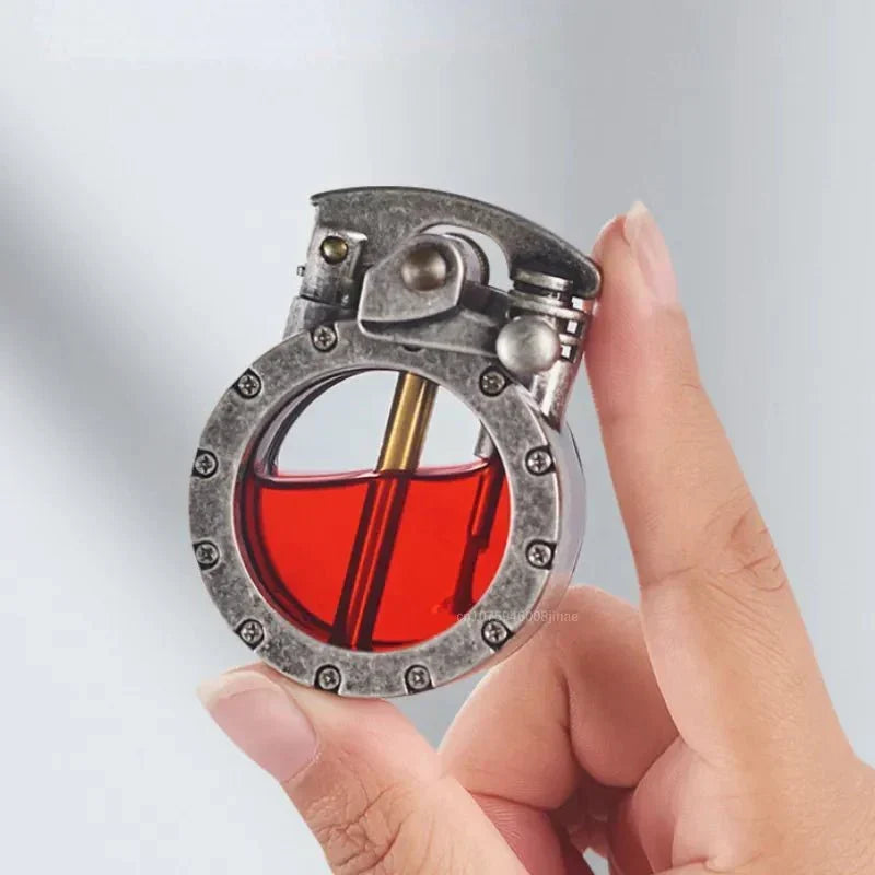 Briquet à cadre métallique vieilli et réservoir transparent rouge tenu entre deux doigts, montrant sa petite taille. 2
