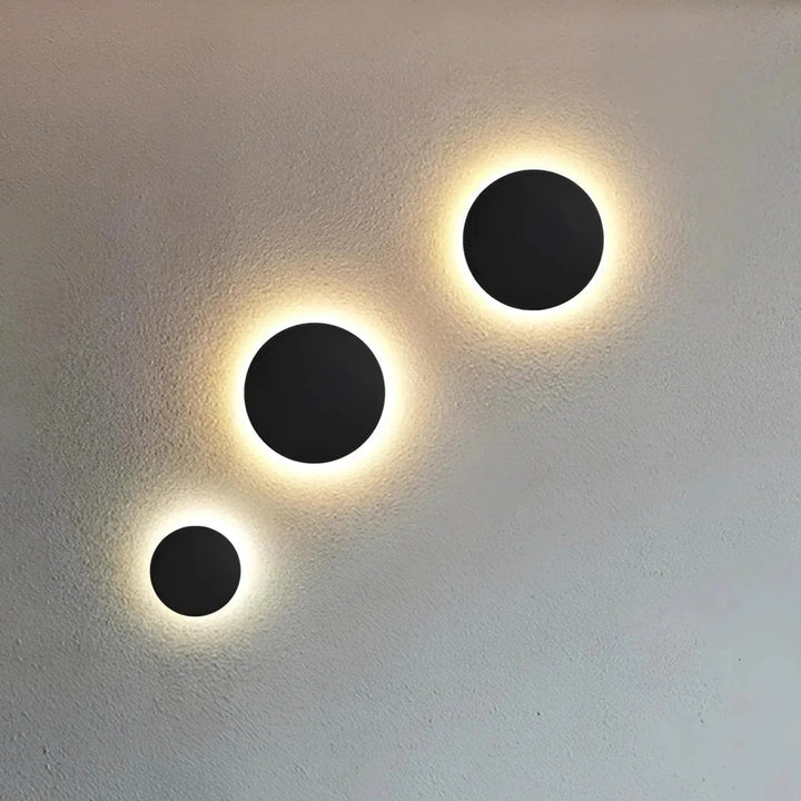 Trois appliques rondes noires de tailles différentes, alignées en diagonale sur un mur texturé, avec un halo lumineux chaud. 2