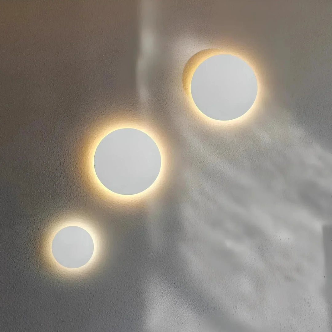 Trois appliques rondes blanches de tailles différentes, disposées en diagonale sur un mur gris, diffusant une lumière chaude en halo. 4