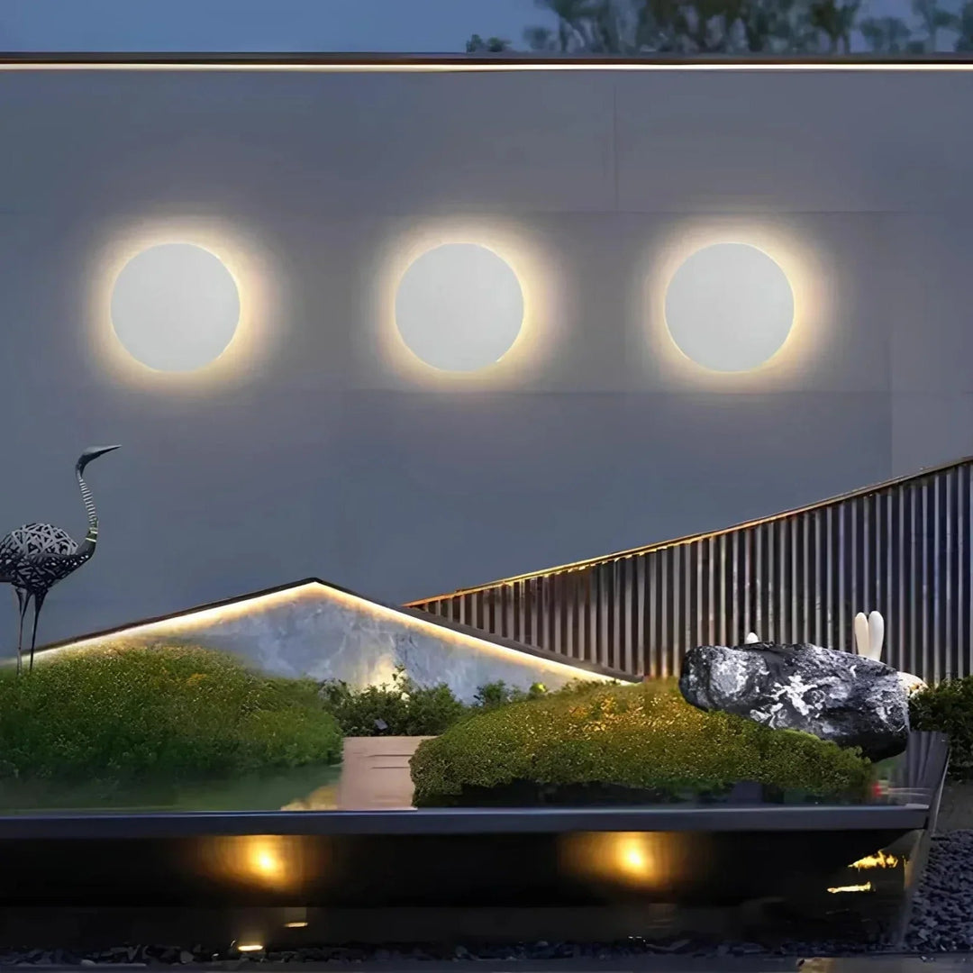 Trois appliques rondes blanches alignées sur un mur extérieur au-dessus d'un jardin paysager éclairé, avec rochers et mousse. 5