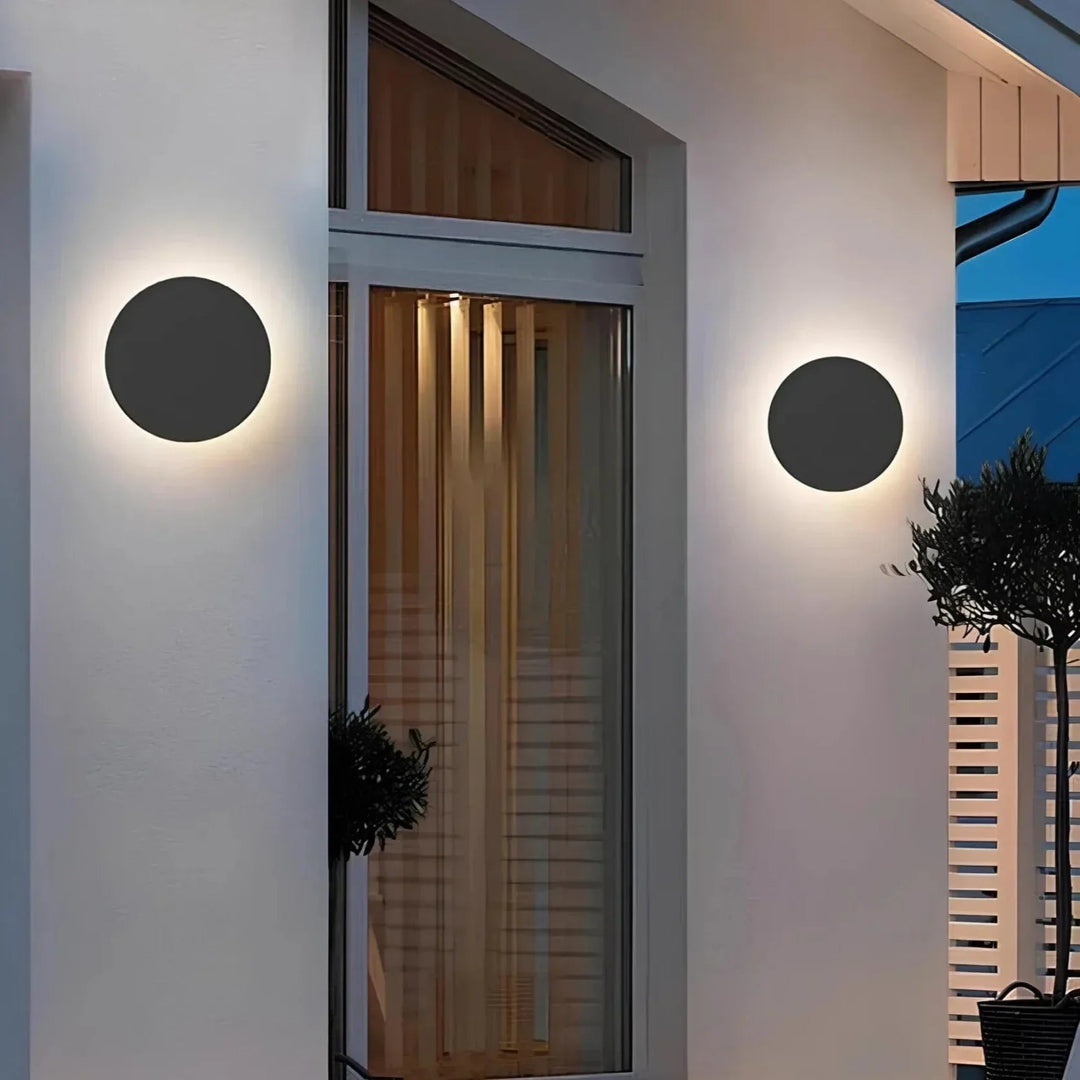 Deux appliques rondes noires fixées sur la façade blanche d'une maison, de part et d'autre d'une grande fenêtre, au crépuscule. 3