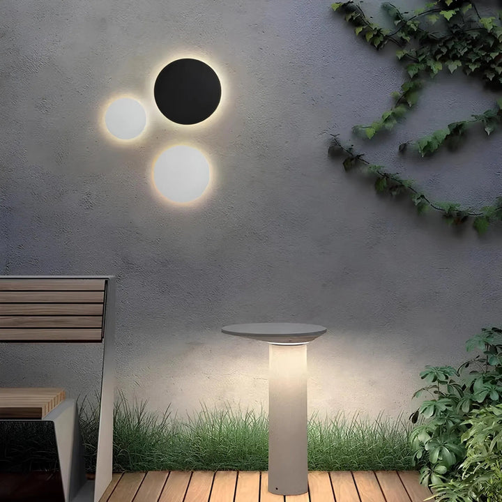 Trois appliques rondes, une noire et deux blanches, éclairant un mur de patio à côté d'une borne lumineuse et d'un banc. 1
