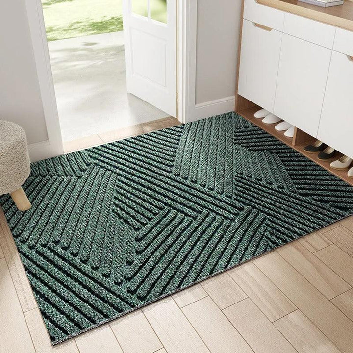Paillasson vert foncé à rayures en relief formant des triangles géométriques, posé dans une entrée devant un meuble à chaussures. 1