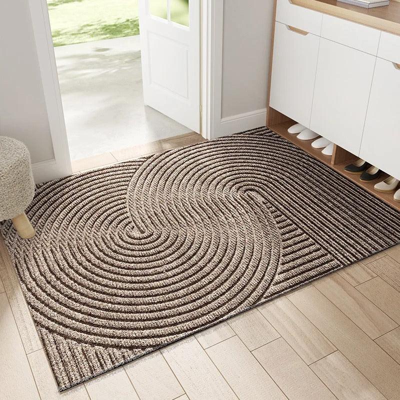 Paillasson beige-brun à cercles concentriques entrelacés en relief, posé sur un parquet clair dans l'entrée d'une maison. 3