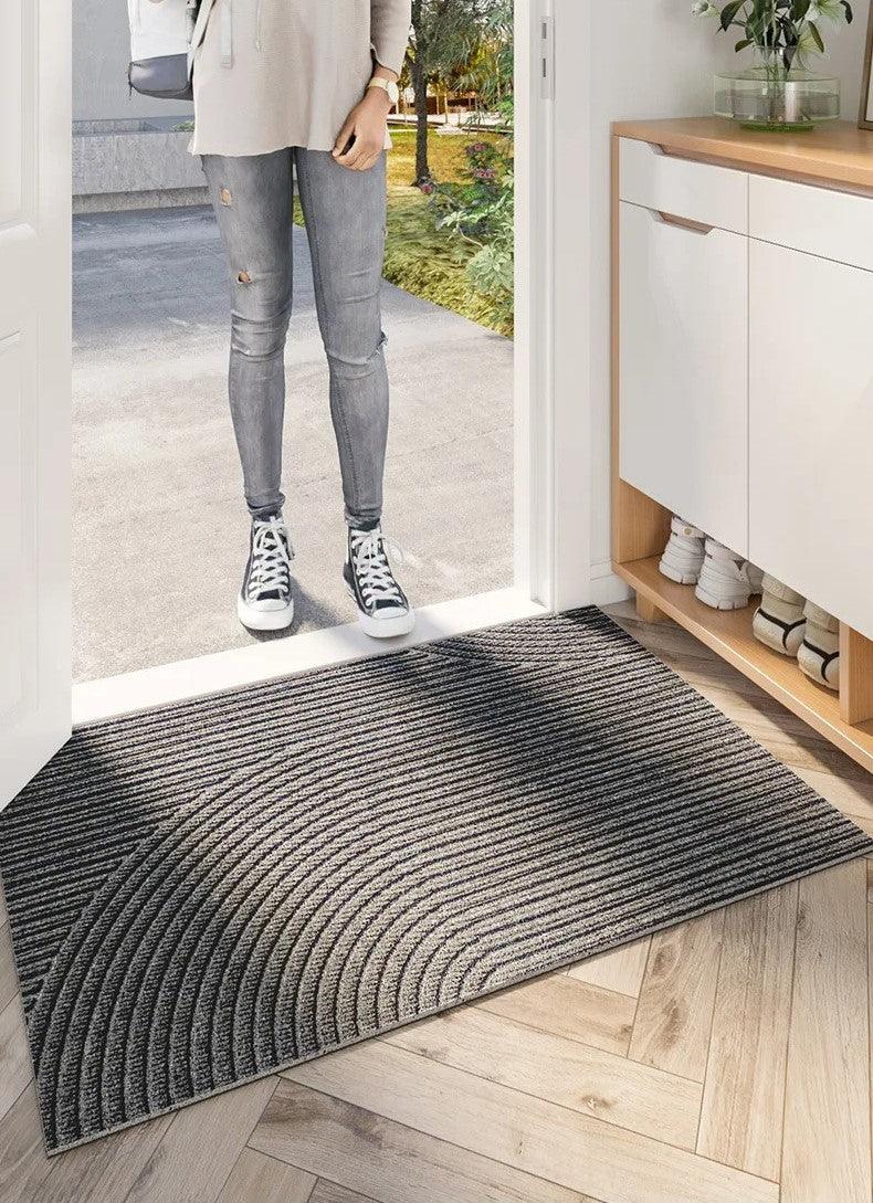 Paillasson gris à rayures courbes en relief devant une porte ouverte, avec une personne en jean et baskets sur le seuil. 5