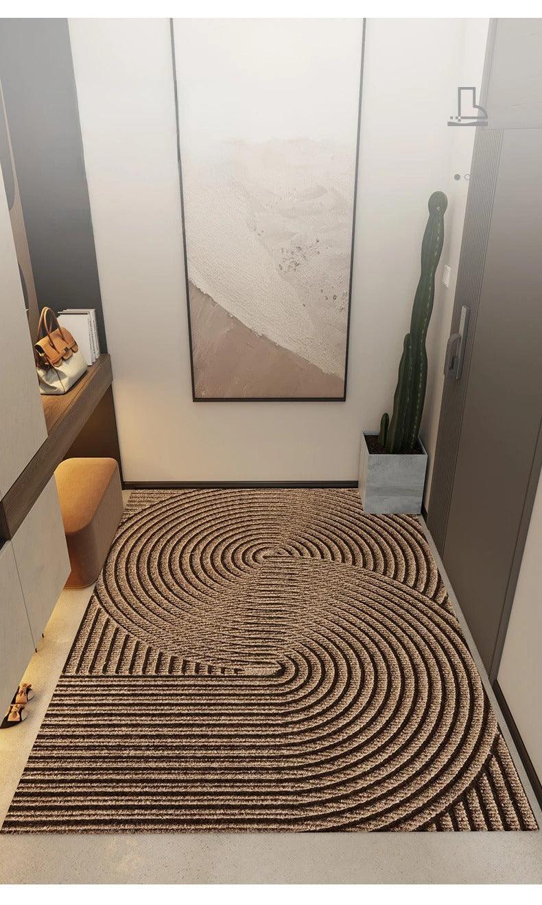 Paillasson brun à grands cercles concentriques en relief, posé dans un couloir d'entrée moderne avec cactus, tableau et pouf. 6