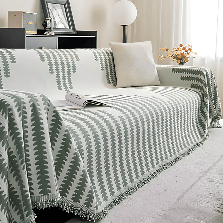 Version vert sauge et blanc de la housse sur un canapé, avec magazine ouvert, coussin clair et franges. 10