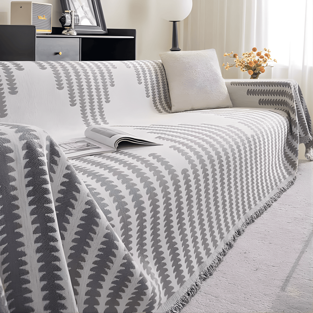 Version grise et blanche de la housse sur un canapé, avec magazine ouvert, coussin clair et franges. 8