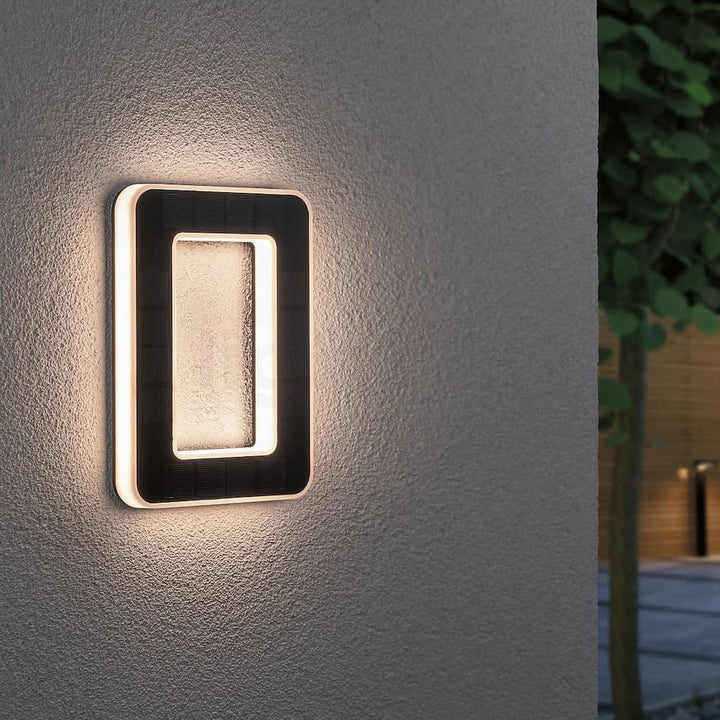 Chiffre 0 lumineux fixé sur un mur crépi gris, cadre noir à panneau solaire et halo de lumière blanc chaud le soir. 1