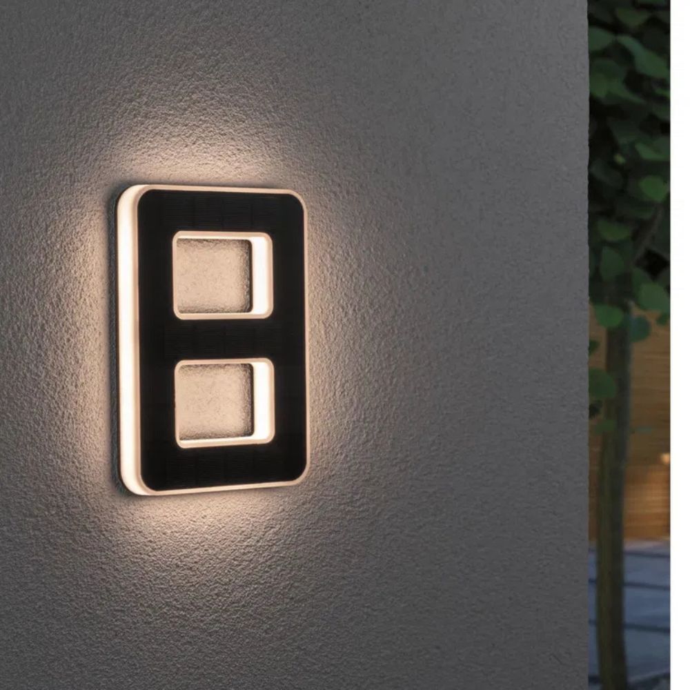 Chiffre 8 lumineux fixé sur un mur crépi, deux ouvertures éclairées et panneau solaire noir entre elles. 9