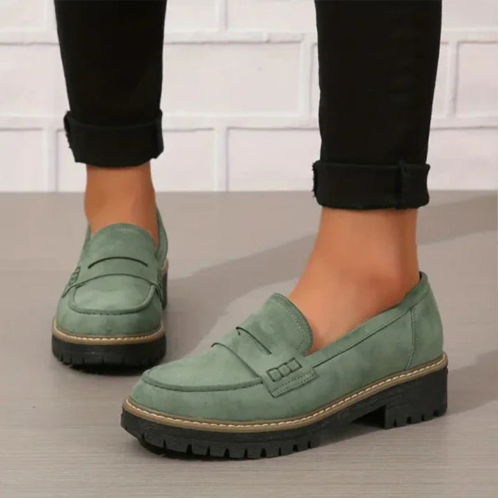 Mocassins verts en daim portés avec un pantalon noir retroussé, vus de trois quarts sur un sol gris devant un mur blanc. 1