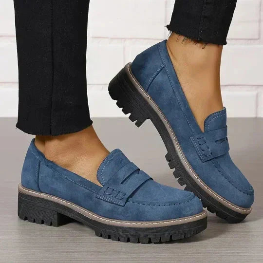 Mocassins bleus portés avec un pantalon noir à l'ourlet effiloché, un pied levé montrant le talon épais. 9