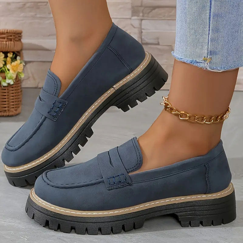 Mocassins bleu marine en daim portés avec une chaîne de cheville dorée, vus de profil sur un sol gris. 16