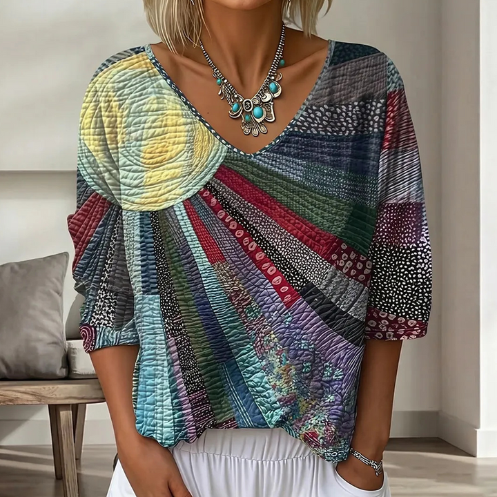 Blouse patchwork à col en V en tons bleu, gris, rouge et vert avec un soleil jaune pâle, portée avec un collier. 5