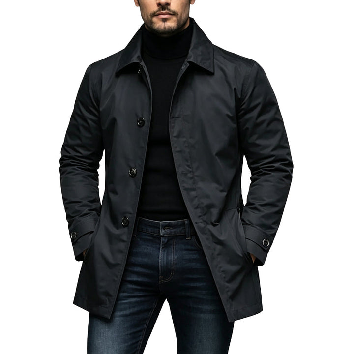 Homme portant le trench noir boutonné, col pointu, avec pull à col roulant noir, jean brut et mains dans les poches. 1