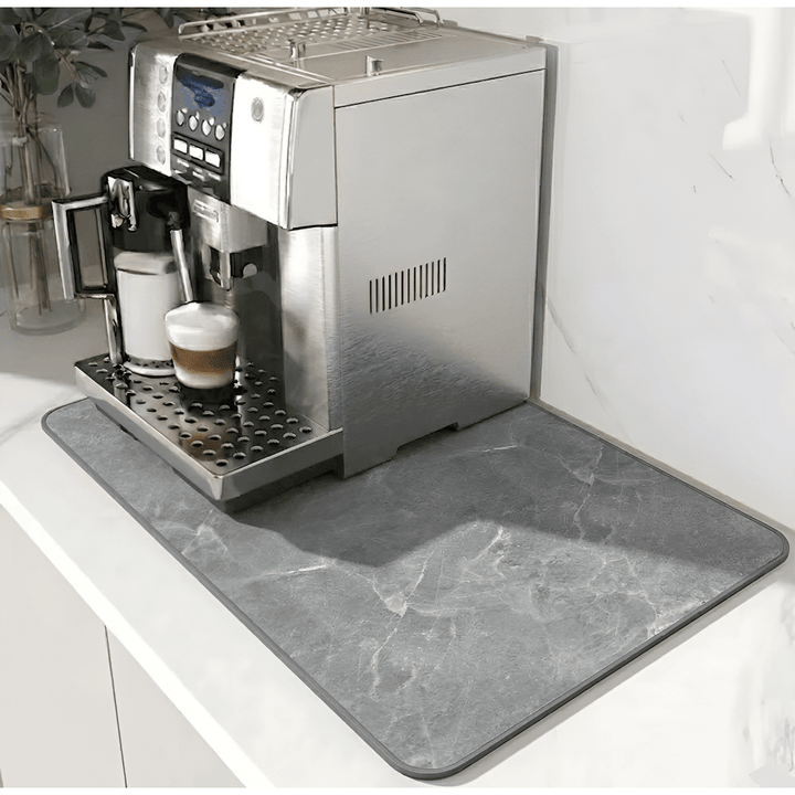 Tapis gris à motif marbré et bordure grise foncée, installé sous une machine à café sur un plan blanc. 12