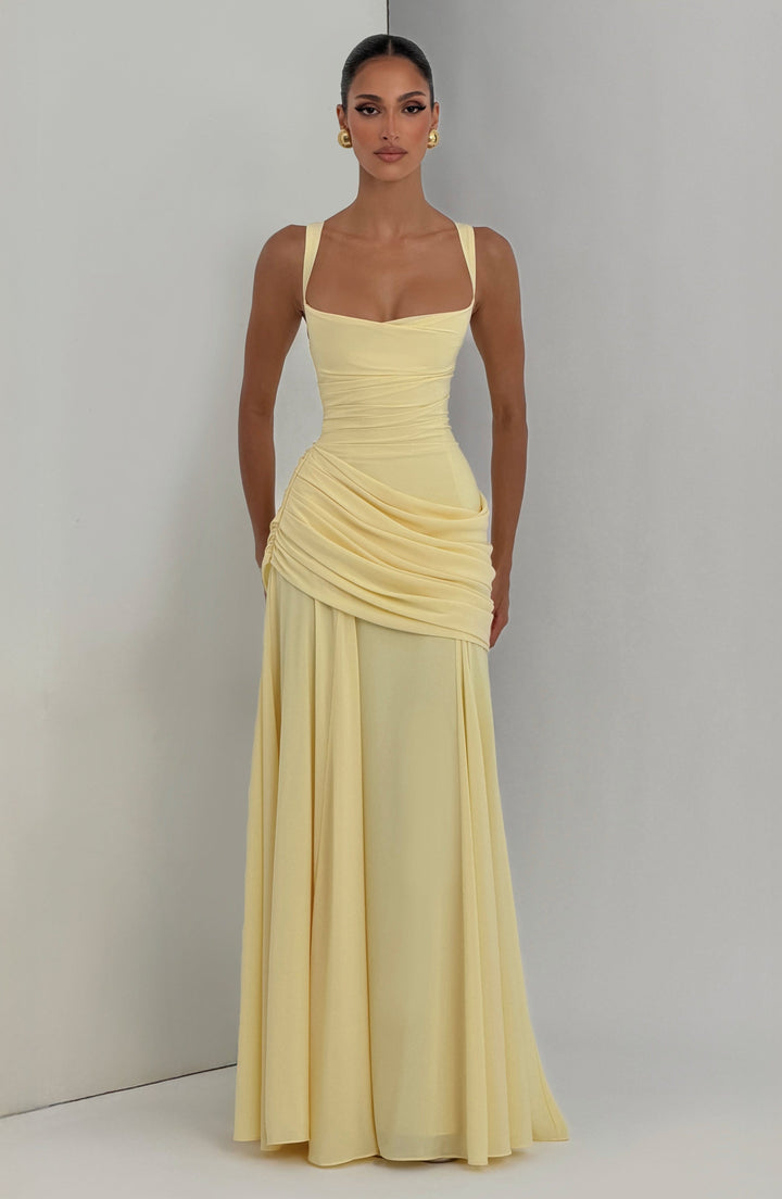 Robe longue jaune pâle portée de face devant un mur gris, jupe évasée et drapé asymétrique sur la hanche. 6