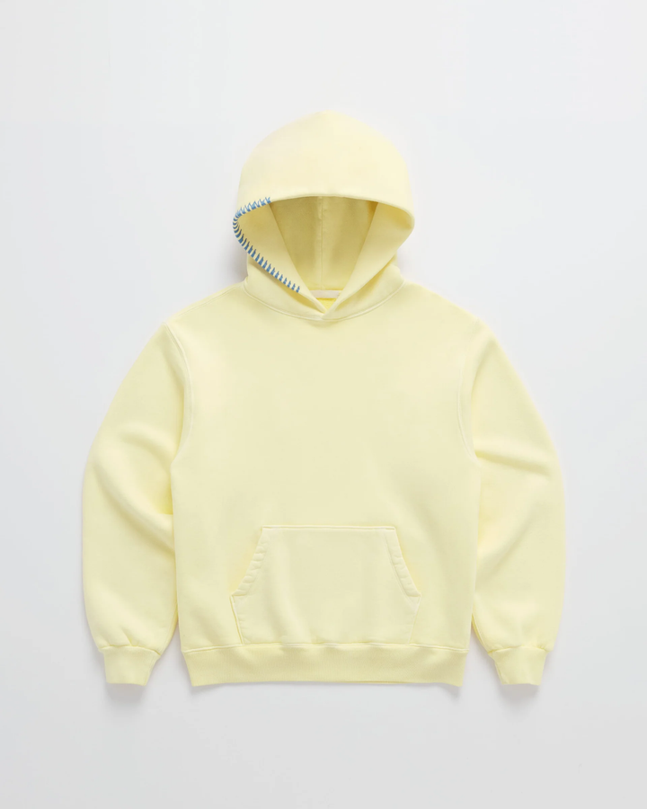 Sweat à capuche jaune pâle posé à plat, poche kangourou ventrale et couture bleue en zigzag sur le bord de la capuche. 1