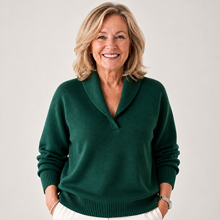 Femme souriante portant le pull vert foncé à col châle, mains dans les poches d'un pantalon blanc, vue de face. 1