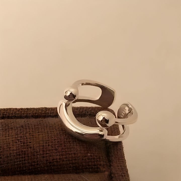 Gros plan sur la bague argentée brillante posée sur un tissu brun tissé, montrant l'ouverture et les deux boules. 5