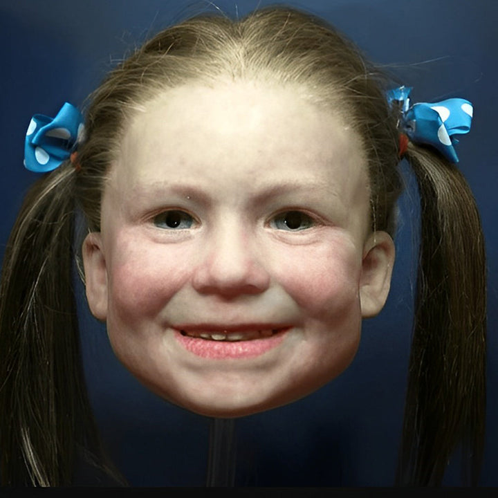 Masque de petite fille souriante aux joues rosées, avec deux couettes brunes nouées par des rubans bleus à pois blancs. 11