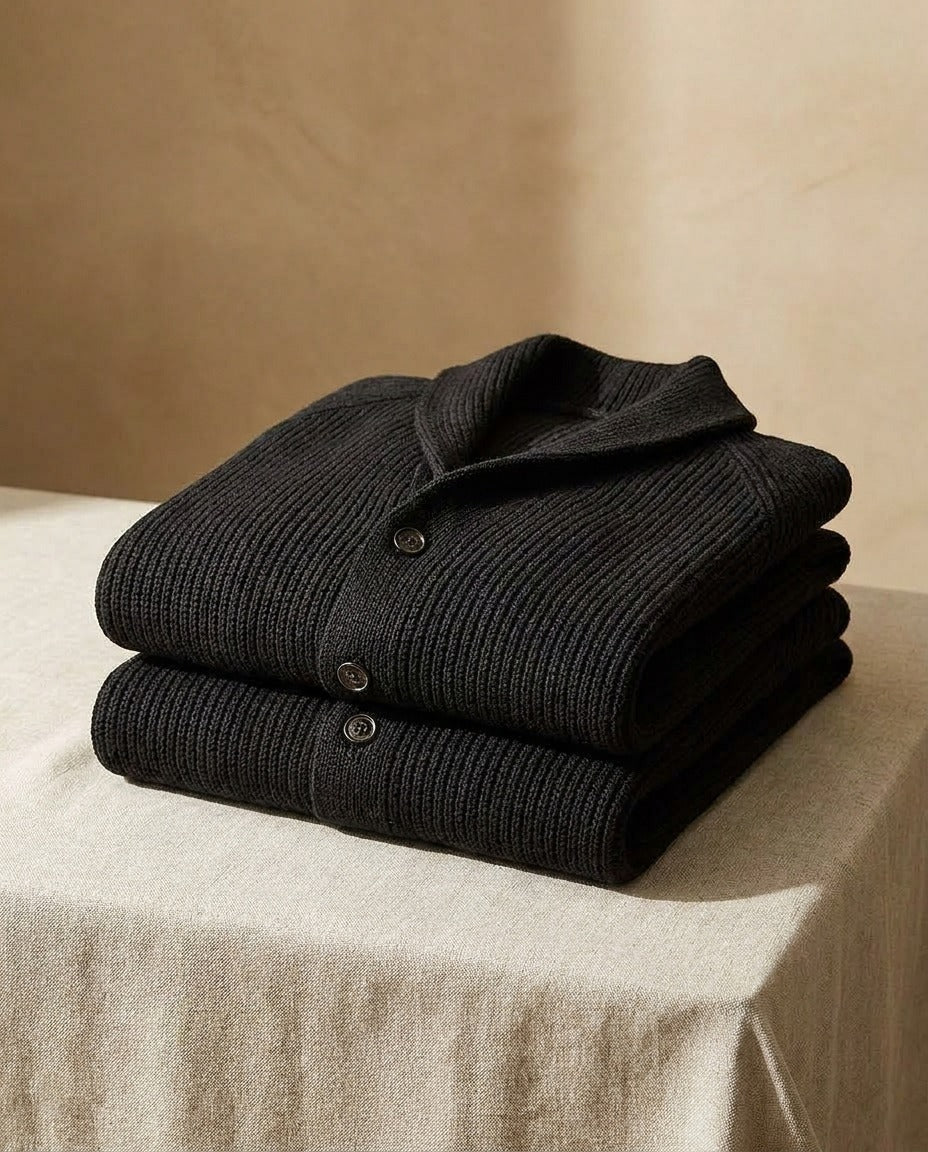 Deux cardigans côtelés noirs pliés et empilés sur une table drapée de lin beige, col châle et boutons visibles. 11