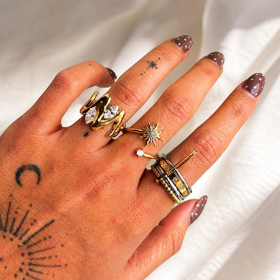 Bague dorée sculptée portée à l'index, superposée à d'autres bagues dorées et argentées, sur une main aux ongles marron. 3