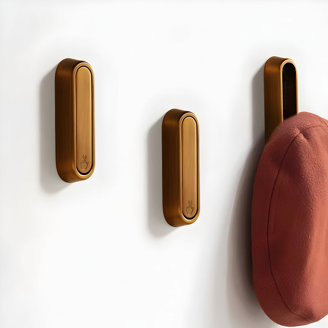 Trois crochets ovales bronze alignés sur un mur blanc, le dernier déplié supportant un bonnet rouge brique. 17