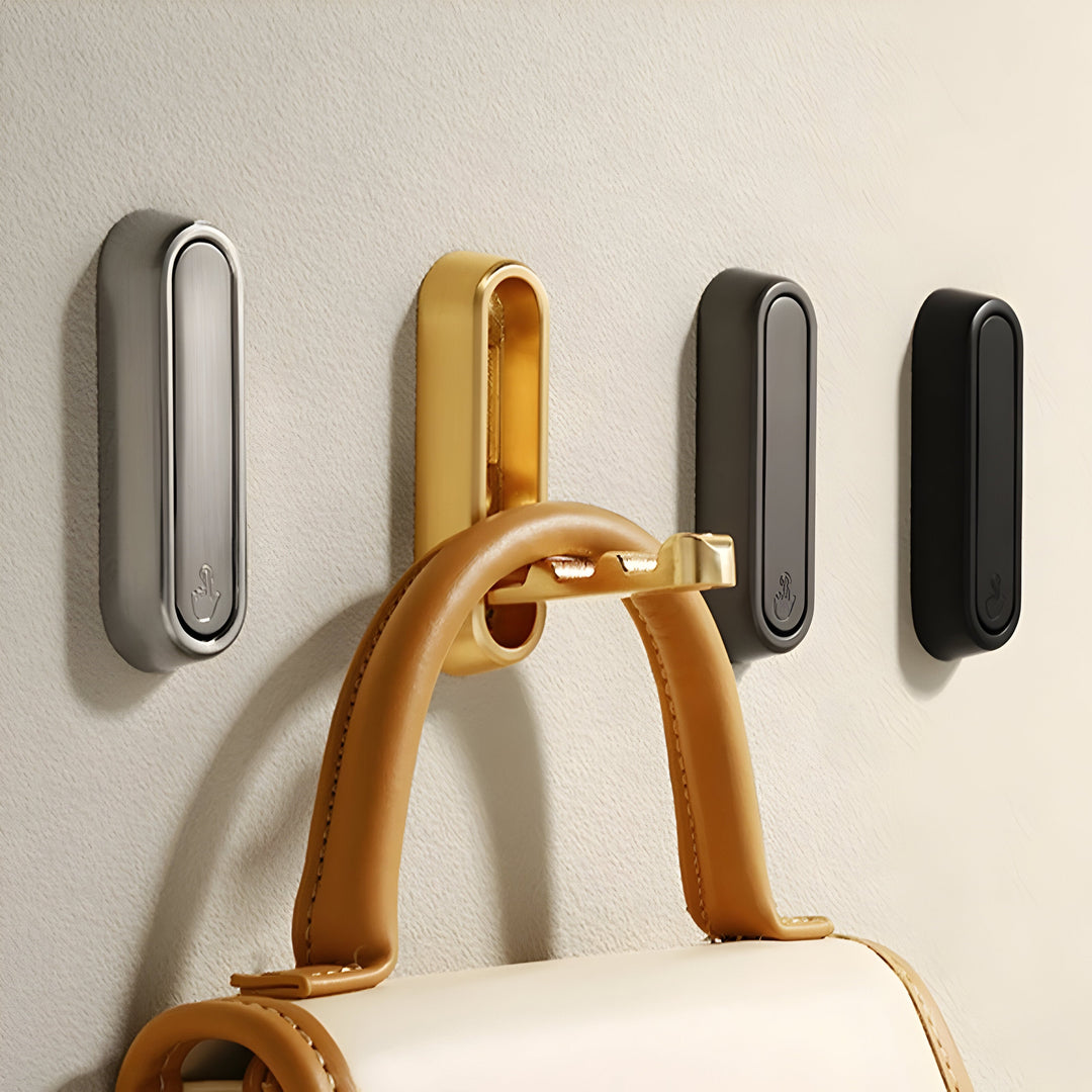 Quatre crochets ovales argenté, doré, gris foncé et noir sur mur clair, le doré déplié portant un sac en cuir. 16