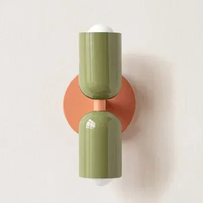 Applique murale à deux tubes laqués vert olive, fixée sur une platine ronde orange corail, sur un mur blanc. 21