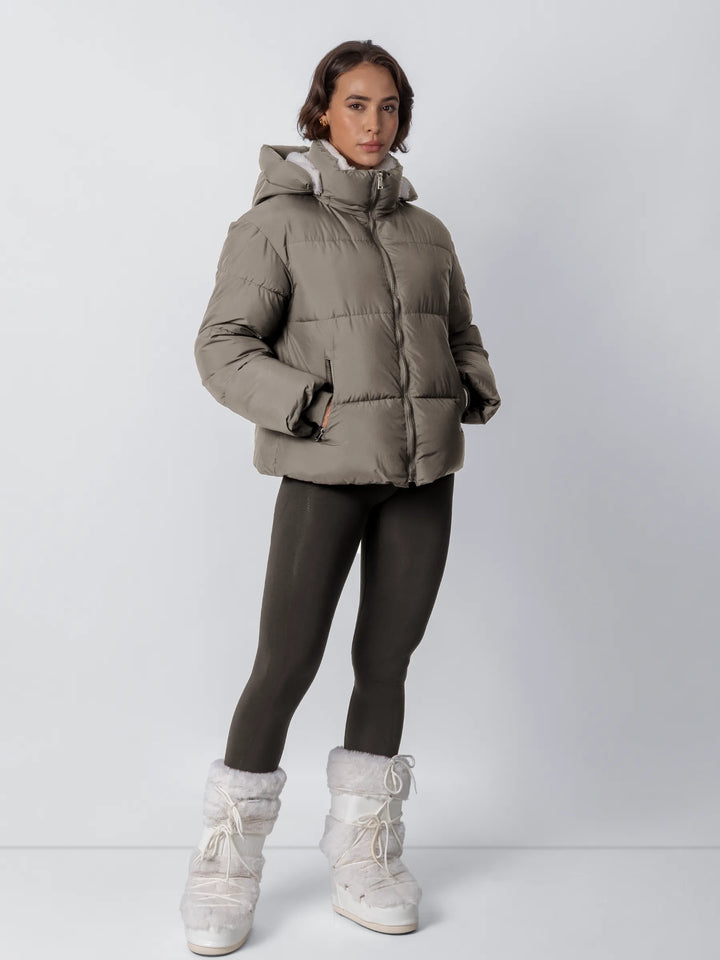 Femme en pied portant la veste kaki zippée, capuche relevée, avec legging foncé et bottes moumoute blanches 21