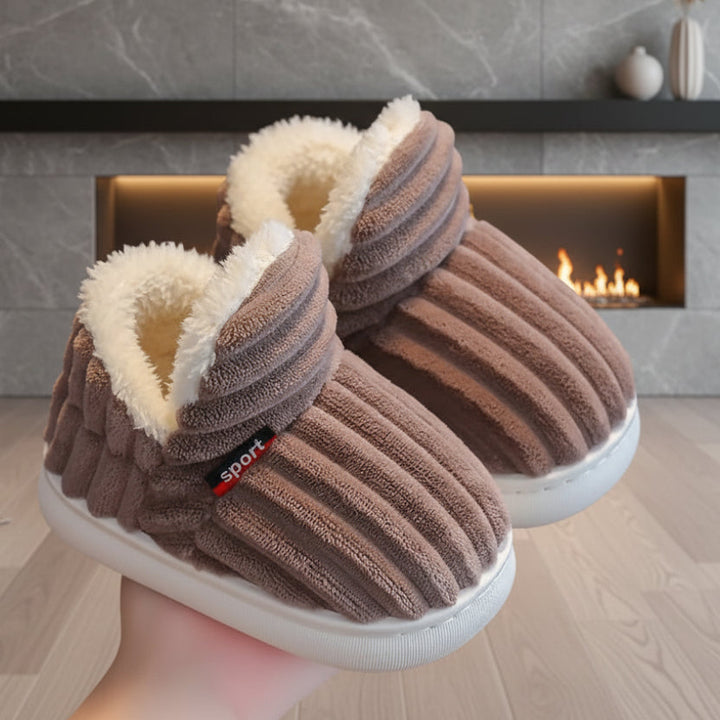 Paire de chaussons marron côtelés à doublure en fausse fourrure blanche, tenue en main devant une cheminée allumée. 1