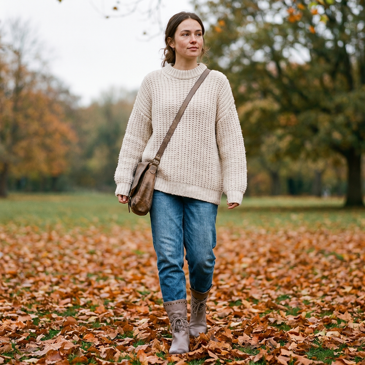 Femme en pull ivoire et jean marchant sur un tapis de feuilles d'automne, avec des bottes grises lacées à rivets. 16