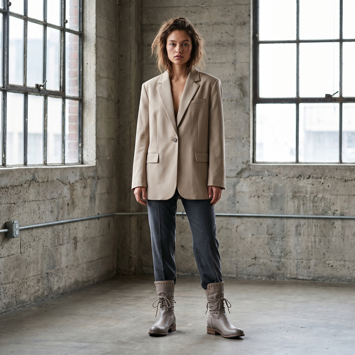 Mannequin en blazer beige et pantalon gris foncé dans un loft au béton brut, portant des bottes grises lacées à rivets. 17