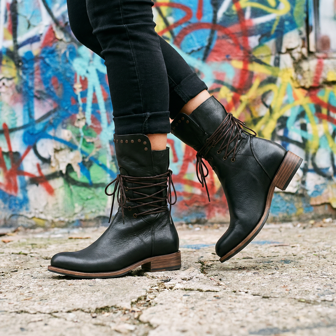 Bottes noires à lacets bruns et rivets portées en marche avec un jean noir retroussé, devant un mur de graffitis colorés. 19