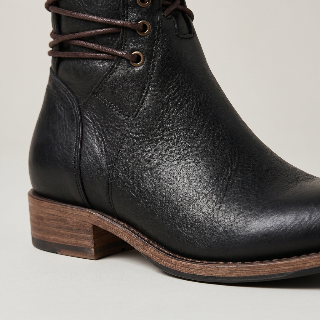 Gros plan sur le bas d'une botte noire: grain du cuir, œillets métalliques, couture de la semelle et talon en bois. 6