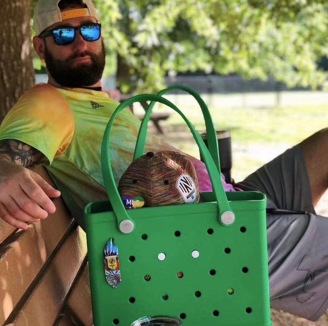 Homme barbu assis sur un banc à l'ombre avec un cabas vert contenant une casquette et orné de petits badges décoratifs. 13