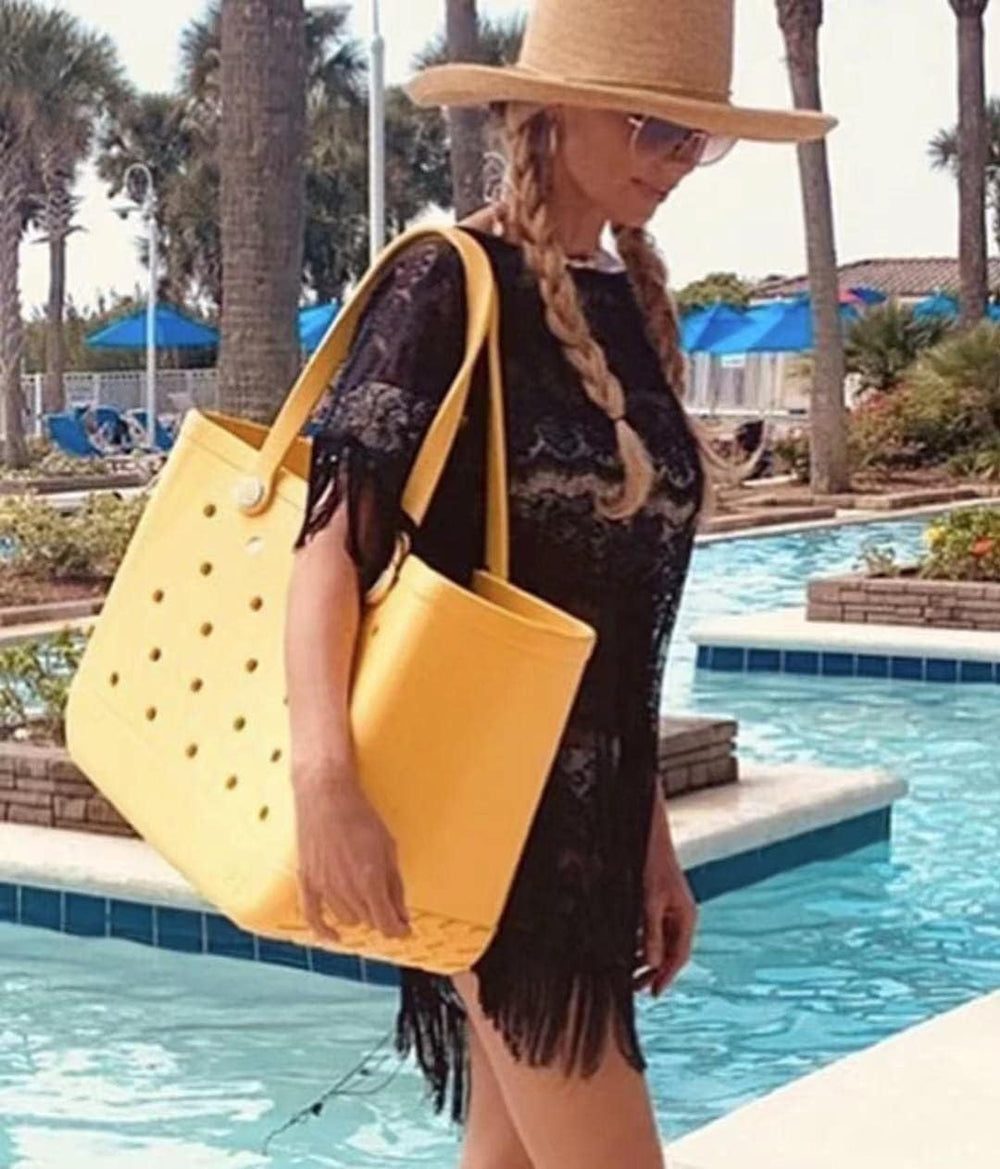Femme en robe de plage noire à franges portant sur l'épaule un grand cabas jaune ajouré au bord d'une piscine. 2
