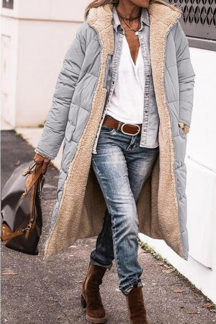 Femme sur un trottoir portant une longue veste matelassée gris clair ouverte, doublure en peluche beige, avec jean et sac à main. 8