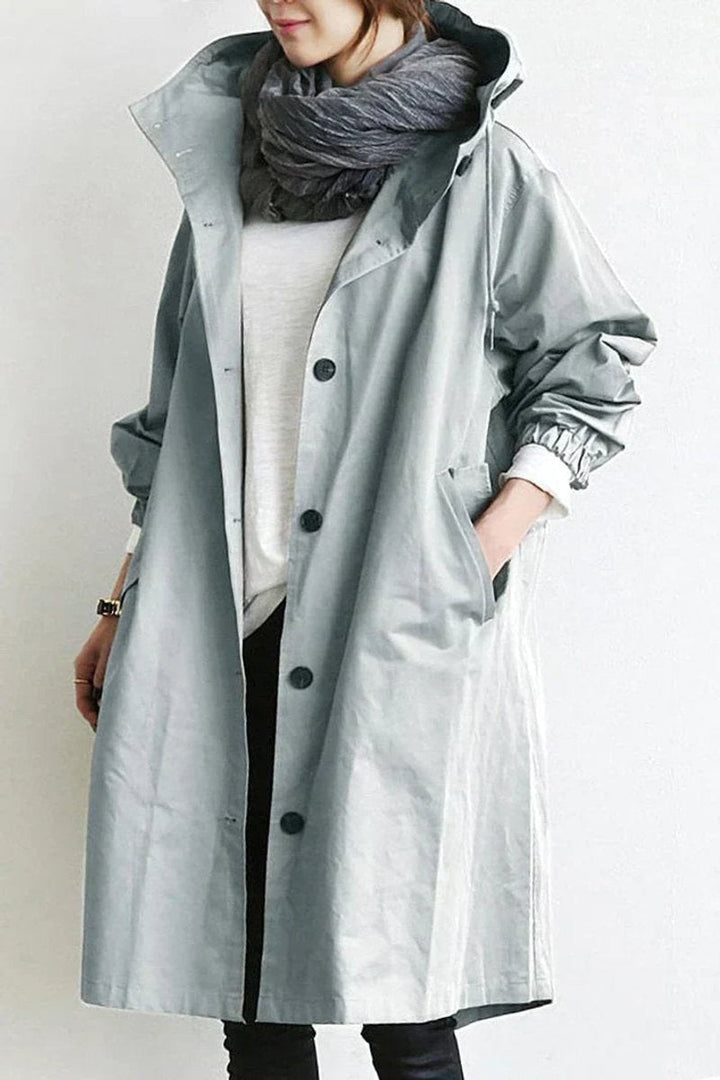 Trench à capuche gris clair boutonné, porté ouvert avec une écharpe grise, une main dans la poche, vue de face. 1