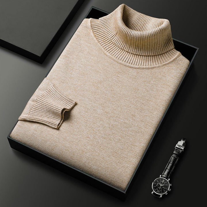 Pull à col roulé beige chiné plié dans une boîte noire, avec manchette côtelée et une montre à bracelet cuir à côté. 1