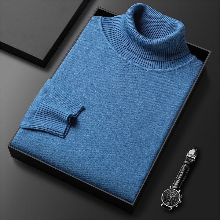 Pull à col roulé bleu plié dans une boîte noire, col et manchette côtelés visibles, montre à bracelet cuir posée à côté. 2