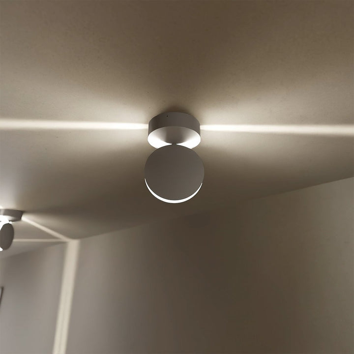 Gros plan sur une applique blanche fixée au plafond, dont la tête ronde diffuse deux traits de lumière horizontaux. 3