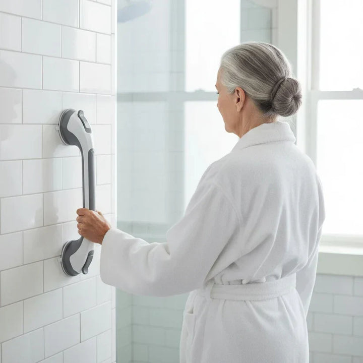 Femme âgée en peignoir blanc, de dos, tenant la barre posée à la verticale sur le carrelage de la douche. 3