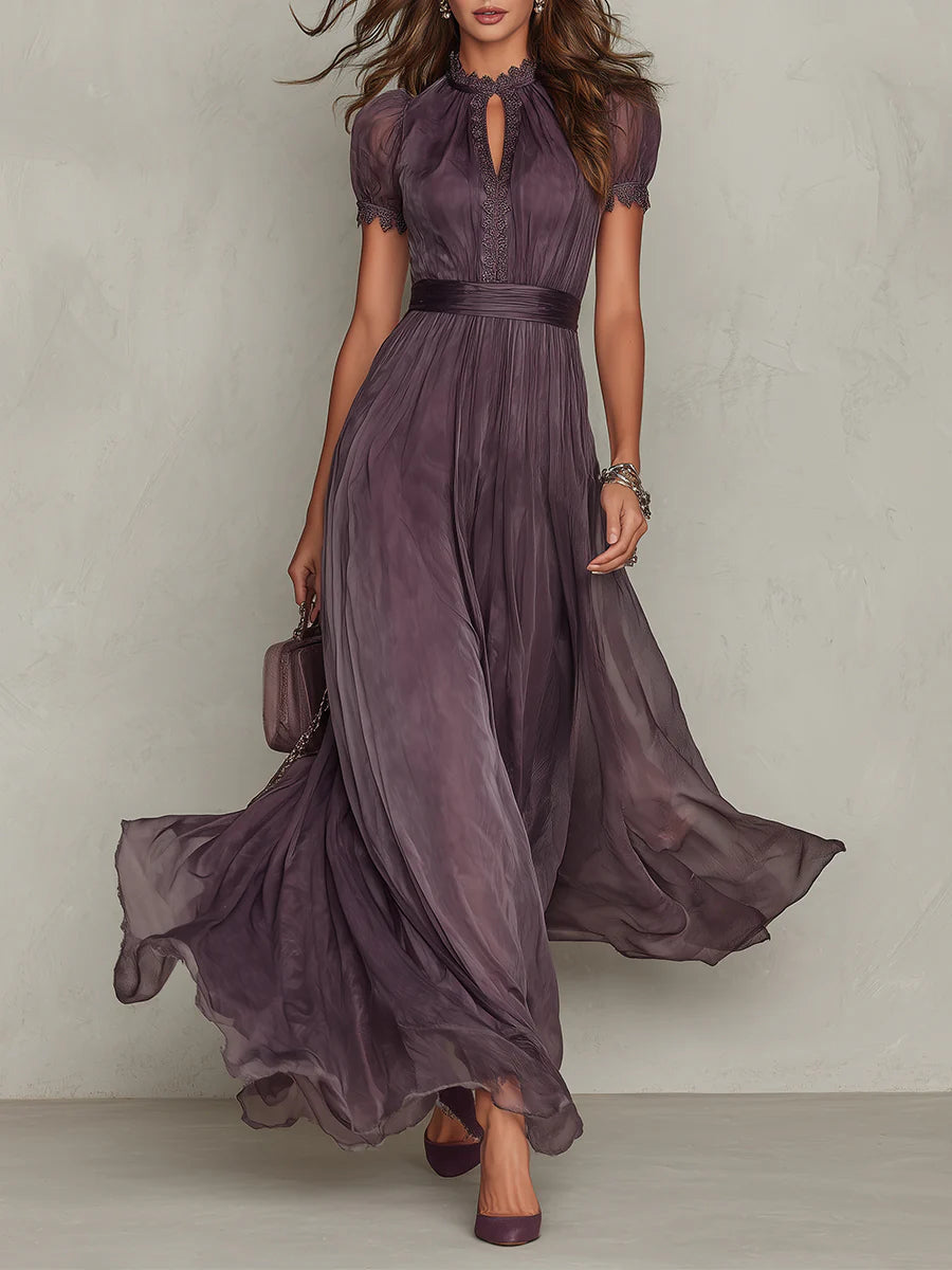Femme en robe longue prune fluide vue de face, avec encolure dentelle, manches bouffantes et jupe ample en mouvement, petit sac à la main. 1