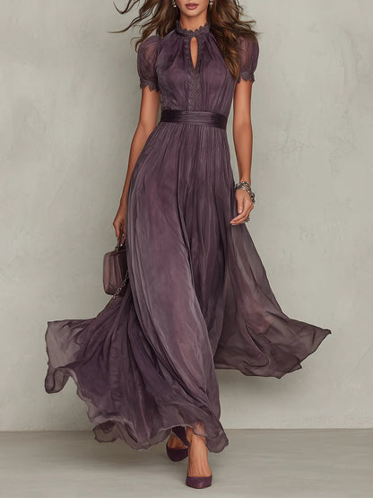 Femme en robe longue prune fluide vue de face, avec encolure dentelle, manches bouffantes et jupe ample en mouvement, petit sac à la main. 1