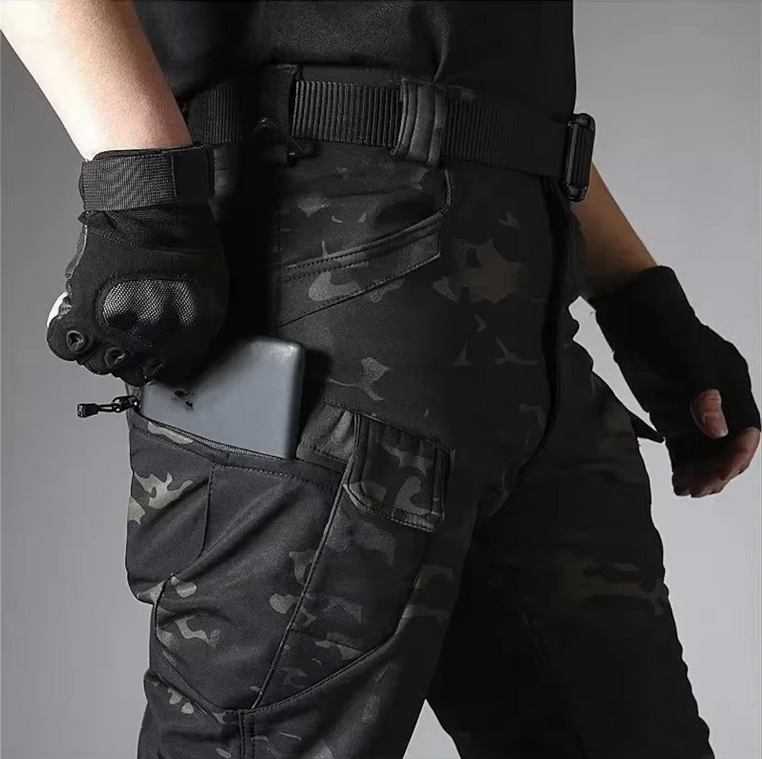 Détail du pantalon camouflage sombre porté avec ceinture et gants noirs, un smartphone glissé dans la poche zippée de la cuisse. 19