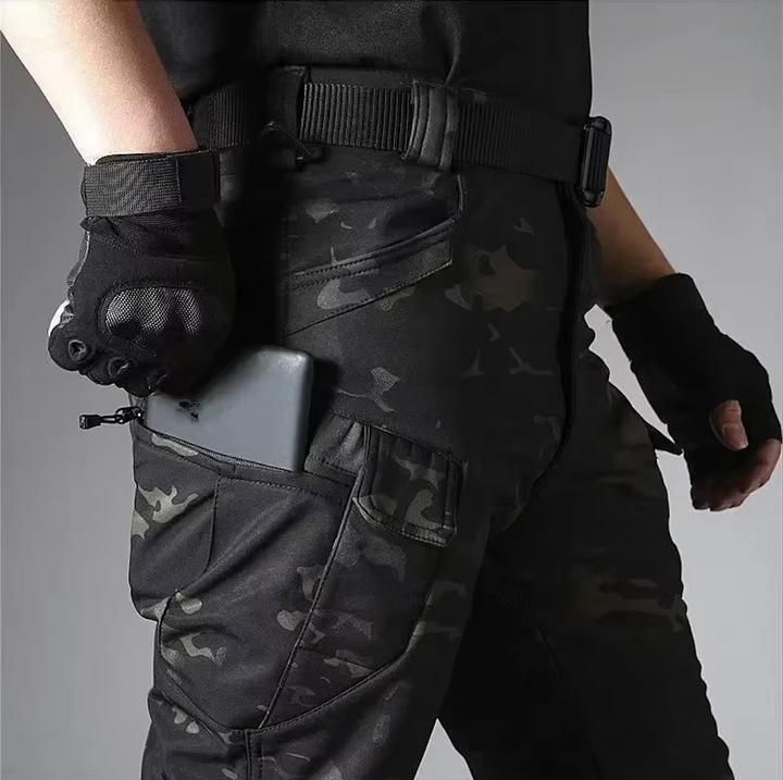 Détail du pantalon camouflage sombre porté avec ceinture et gants noirs, un smartphone glissé dans la poche zippée de la cuisse. 19