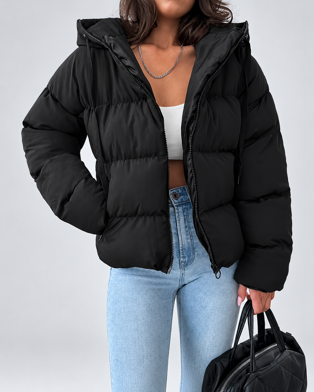 Veste matelassée noire portée ouverte par une femme avec un top blanc, un jean clair et un sac noir à la main. 1